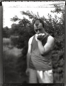 MATTHEW SWARTS A MAN IN A TANK TOP AND SHORTS PLAYS A HARMONICA OUTDOORS, WITH LUSH GREENERY BEHIND HIM.