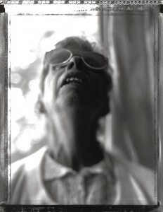MATTHEW SWARTS BLACK AND WHITE VISUAL DOCUMENTARY PHOTO OF AN OLDER PERSON WEARING SUNGLASSES, HEAD TILTED BACK, MOUTH SLIGHTLY OPEN. THE IMAGE IS BLURRED AND OUT OF FOCUS, WITH LIGHT FROM BEHIND CREATING A SOFT, ETHEREAL EFFECT REMINISCENT OF MATTHEW SWARTS’ STYLE.