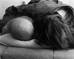 MATTHEW SWARTS A BALD PERSON AND A PERSON WITH LONG HAIR LIE SIDE BY SIDE, HEADS RESTING ON A PILLOW, PHOTOGRAPHED FROM BEHIND IN BLACK AND WHITE—A POIGNANT WORK OF VISUAL ART BY MATTHEW SWARTS THAT QUIETLY EVOKES THE PRESENCE OF CANCER.