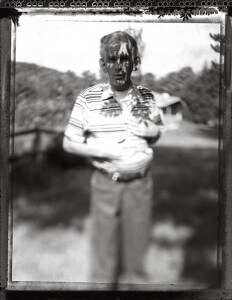 MATTHEW SWARTS A BLACK-AND-WHITE, BLURRED VISUAL CAPTURES A MAN STANDING OUTDOORS IN SUNLIGHT. SHADOWS OF LEAVES CROSS HIS FACE. HE WEARS A STRIPED SHIRT AND HOLDS HIS HAND TO HIS CHEST, WITH TREES AND A BUILDING BEHIND—A CONTEMPORARY WORK BY MATTHEW SWARTS.