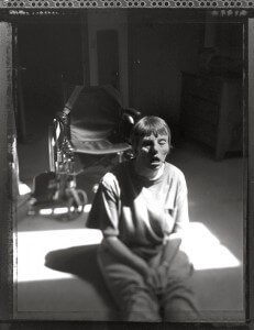 MATTHEW SWARTS A PERSON SITS ON THE FLOOR IN A PATCH OF SUNLIGHT, EYES CLOSED, WITH A WHEELCHAIR POSITIONED IN THE BACKGROUND AND A DRESSER NEARBY IN A DIMLY LIT ROOM—AN INTIMATE MOMENT CAPTURED THROUGH THE LENS OF DOCUMENTARY PHOTOGRAPHY BY MATTHEW SWARTS.