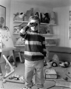 MATTHEW SWARTS A BLACK AND WHITE PHOTOGRAPHY CAPTURES A YOUNG CHILD IN A STRIPED SWEATER PEERING THROUGH BINOCULARS IN A PLAYROOM, SURROUNDED BY TOYS AND SHELVES OF STUFFED ANIMALS—A TOUCHING MOMENT REFLECTING THE WORLD OF CHILDREN WITH CANCER.