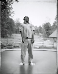 MATTHEW SWARTS A PERSON STANDS BAREFOOT OUTDOORS ON A TRAMPOLINE, SMILING AND LOOKING UP IN THIS CONTEMPORARY BLACK AND WHITE IMAGE. WEARING A SHORT-SLEEVE POLO SHIRT AND PANTS, THEY APPEAR CANDID AS BLURRED TREES AND A BUILDING EVOKE A DOCUMENTARY FEEL IN THE BACKGROUND.