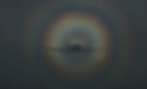 MATTHEW SWARTS A BLURRY, DARK IMAGE SHOWING A CIRCULAR RAINBOW-LIKE HALO WITH ORANGE AND BLUE HUES SURROUNDING A DARK CENTRAL SHAPE, SET AGAINST A GREY, OVERCAST SKY.