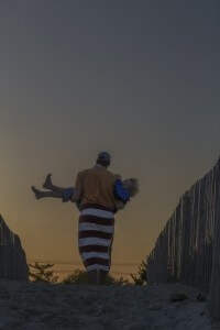 MATTHEW SWARTS A PERSON CARRYING A CHILD, BOTH SILHOUETTED AGAINST A DUSKY SKY. A STRIPED TOWEL WRAPS AROUND THE PERSONS LEGS.