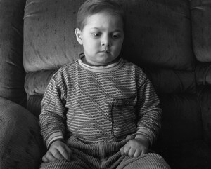 MATTHEW SWARTS A YOUNG CHILD WITH SHORT HAIR SITS ON A CUSHIONED CHAIR, LOOKING DOWN WITH A SERIOUS EXPRESSION. THE CHILD WEARS STRIPED LONG-SLEEVE PAJAMAS. THIS BLACK AND WHITE PHOTOGRAPH REFLECTS THE SIGNATURE STYLE OF MATTHEW SWARTS’S VISUAL ART.