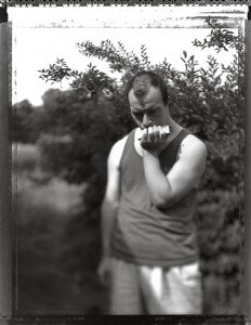 MATTHEW SWARTS A MAN IN A SLEEVELESS SHIRT STANDS OUTDOORS IN FRONT OF LEAFY BUSHES, LOOKING DOWN THOUGHTFULLY WHILE HOLDING A SMALL OBJECT TO HIS FACE. THE BLACK-AND-WHITE, SLIGHTLY OUT-OF-FOCUS PHOTOGRAPHY GIVES THE SCENE A CONTEMPORARY FEEL.