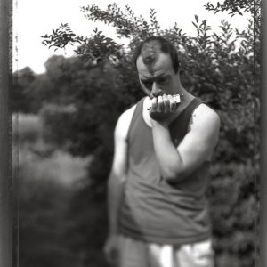 MATTHEW SWARTS A MAN IN A SLEEVELESS SHIRT STANDS OUTDOORS IN FRONT OF LEAFY BUSHES, LOOKING DOWN THOUGHTFULLY WHILE HOLDING A SMALL OBJECT TO HIS FACE. THE BLACK-AND-WHITE, SLIGHTLY OUT-OF-FOCUS PHOTOGRAPHY GIVES THE SCENE A CONTEMPORARY FEEL.