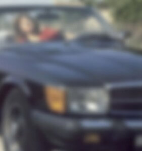 MATTHEW SWARTS A PERSON SITTING IN THE DRIVER’S SEAT OF A BLACK CONVERTIBLE CAR, OUTDOORS ON A SUNNY DAY. THE IMAGE IS BLURRY, MAKING FACIAL DETAILS AND BACKGROUND DIFFICULT TO SEE.