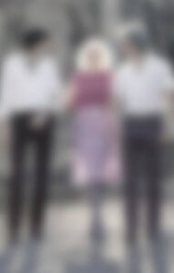 MATTHEW SWARTS THREE PEOPLE STAND SIDE BY SIDE OUTDOORS. THE PERSON IN THE MIDDLE WEARS A PINK TOP AND PURPLE SKIRT, WHILE THE OTHERS ON EITHER SIDE WEAR WHITE TOPS AND DARK PANTS. THE BACKGROUND APPEARS TO BE NATURAL OR ROCKY.