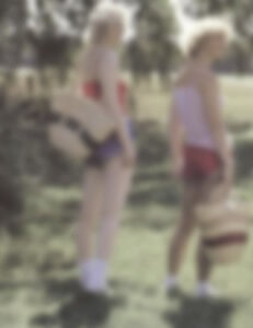 MATTHEW SWARTS TWO PEOPLE STANDING OUTDOORS ON GRASS, BOTH WEARING SHORTS, SLEEVELESS TOPS, AND HOLDING HATS. THE IMAGE IS VERY BLURRY, MAKING FACES AND DETAILS INDISTINGUISHABLE. TREES AND SUNLIGHT ARE VISIBLE IN THE BACKGROUND.