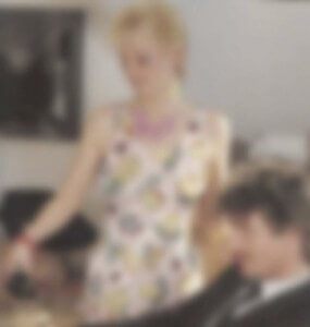 MATTHEW SWARTS A BLURRED IMAGE OF A WOMAN IN A FLORAL DRESS STANDING NEXT TO A SEATED MAN IN A SUIT, INDOORS, WITH INDISTINCT BACKGROUND DETAILS.