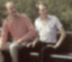 MATTHEW SWARTS TWO MEN SIT ON A BENCH OUTDOORS, WITH GREENERY IN THE BACKGROUND. THE IMAGE IS VERY BLURRY, MAKING FACIAL FEATURES AND DETAILS OF THEIR CLOTHING INDISTINGUISHABLE.