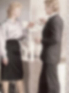 MATTHEW SWARTS TWO PEOPLE STANDING BY A WATER COOLER, ONE WEARING A SUIT AND THE OTHER IN A BLOUSE AND SKIRT, APPEAR TO BE HAVING A CONVERSATION. THE IMAGE IS VERY BLURRY, SO FACIAL FEATURES AND DETAILS ARE UNCLEAR.