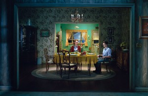 MATTHEW SWARTS A WOMAN AND A TEENAGE BOY SIT AT A DINING TABLE COVERED WITH A YELLOW CLOTH IN A DIMLY LIT, VINTAGE-STYLE ROOM. THE TABLE IS SET FOR A MEAL. THE BOY SITS APART FROM THE WOMAN, BOTH APPEARING SERIOUS AND DISTANT.