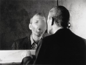 MATTHEW SWARTS BLACK AND WHITE PHOTO OF A MAN LOOKING INTO A MIRROR. HIS FACE IN THE REFLECTION IS BLURRED. HE IS WEARING A DARK JACKET AND SHIRT.
