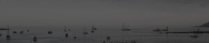 MATTHEW SWARTS A DARK, MOODY BLACK-AND-WHITE PANORAMIC VIEW OF A CALM HARBOR WITH NUMEROUS SAILBOATS AND YACHTS ANCHORED ON STILL WATER UNDER AN OVERCAST SKY.