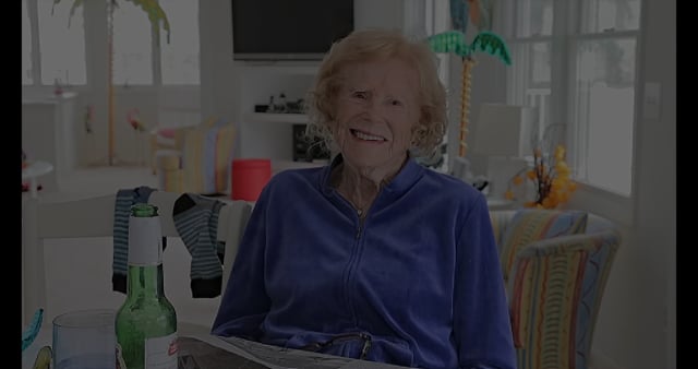 MATTHEW SWARTS An elderly woman with light hair sits smiling at a table in a bright, sunlit room with colorful chairs and contemporary decor. A green bottle, glass, and newspaper rest before her—perfect for a moment of candid photography.