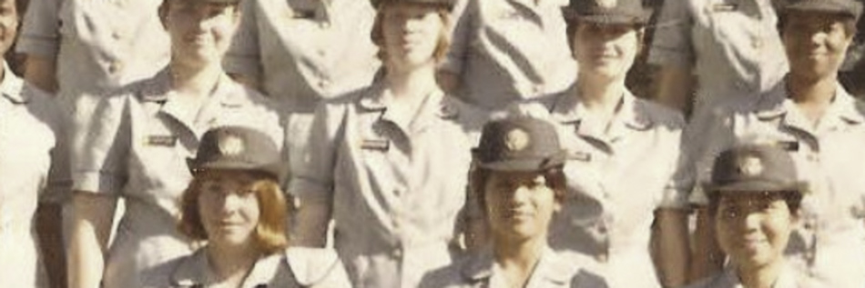 MATTHEW SWARTS A group of women in uniform, standing in rows and smiling. They are wearing light-colored shirts and matching hats with emblems, posing for a group photo outdoors like a scene from contemporary art.