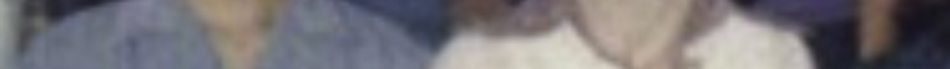 MATTHEW SWARTS A very blurry image in the style of fine art photography shows two people standing side by side, one in a blue top and the other in white. Their faces and background details are indistinguishable, emphasizing mood over clarity.