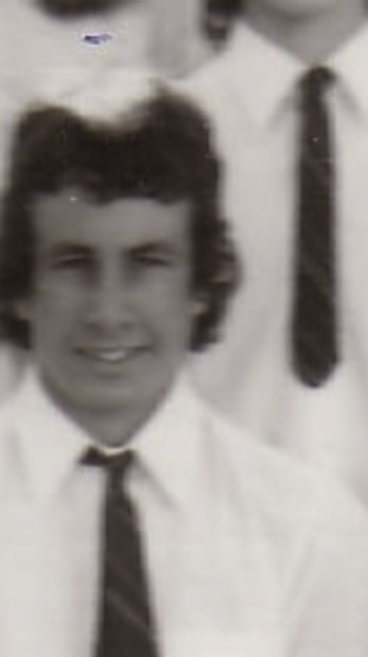 MATTHEW SWARTS A black-and-white photo in the style of fine art photography shows a young person with wavy dark hair, wearing a white shirt and dark tie, smiling at the camera. The blurred background features another person partly visible.