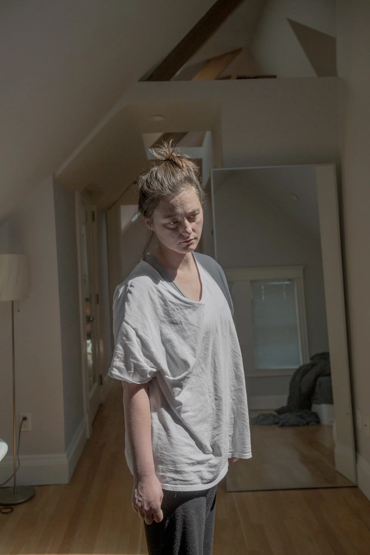 MATTHEW SWARTS A young woman with a messy bun stands in a sunlit, contemporary room, wearing a loose white T-shirt and dark pants, looking downward with a tired expression. Light streams through a window, and a large mirror stands behind her.