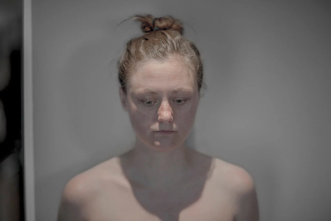 MATTHEW SWARTS A woman with fair skin and reddish hair tied in a messy bun looks down with a neutral expression, standing against a plain gray background—a striking visual reminiscent of artist Matthew Swarts' style. She appears to be unclothed from the shoulders up.
