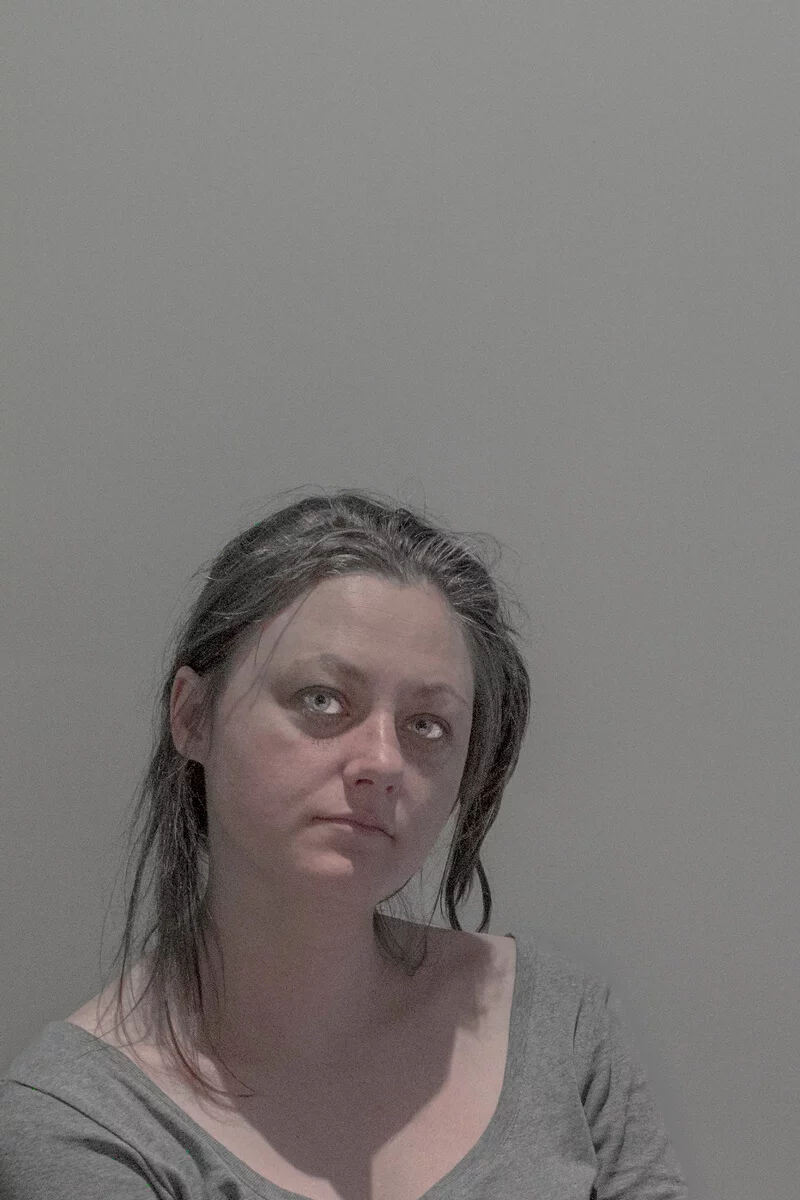 MATTHEW SWARTS A woman with pale skin and dark, slightly messy hair sits against a plain gray background, wearing a gray top. She looks slightly upward with a neutral expression—a quiet moment captured in the ART of MATTHEW SWARTS. The lighting is soft and even.