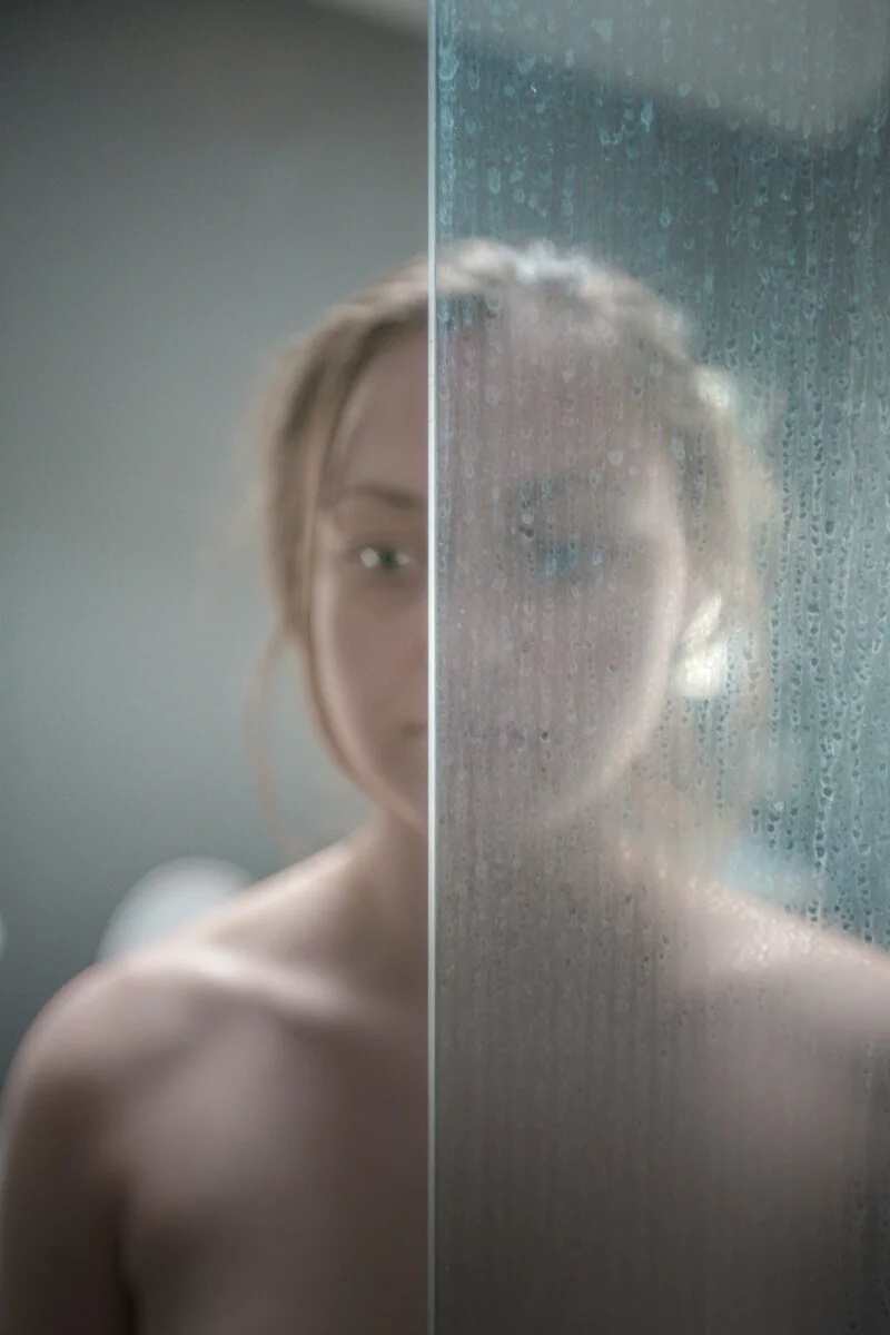 MATTHEW SWARTS A person stands behind a glass panel, one half clear and the other covered in water droplets, blurring part of their face and body. The background is softly out of focus in this evocative image by photographer Matthew Swarts.