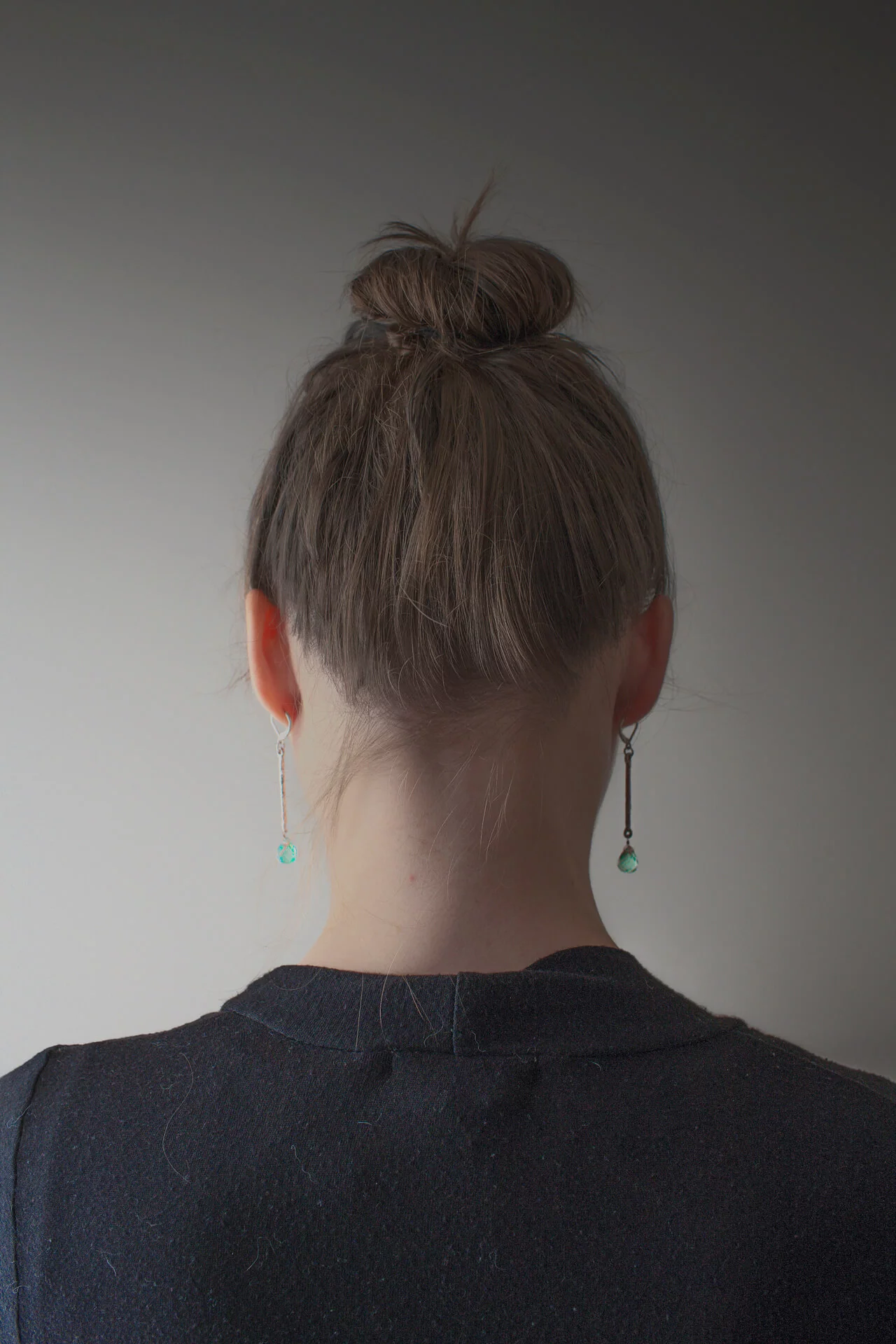 MATTHEW SWARTS A person with light brown hair in a messy bun, wearing a dark top and long earrings with green pendants, is shown from behind against a plain, softly lit background, evoking a contemporary VISUAL artist’s portrait.