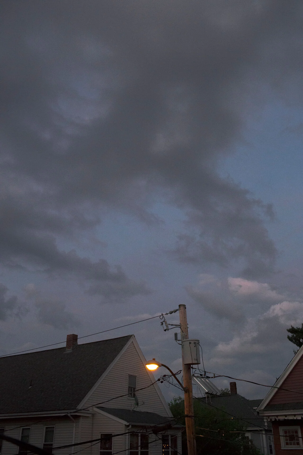 MATTHEW SWARTS A contemporary photograph captures a streetlight glowing near a utility pole in front of two houses with pitched roofs under a cloudy evening sky.