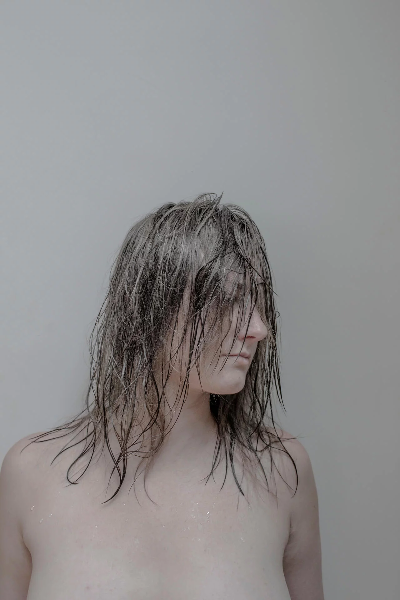 MATTHEW SWARTS A person with wet, shoulder-length hair stands against a plain, light background, looking to the side. Their hair partially covers their face, and their bare shoulders evoke a contemporary art portrait.