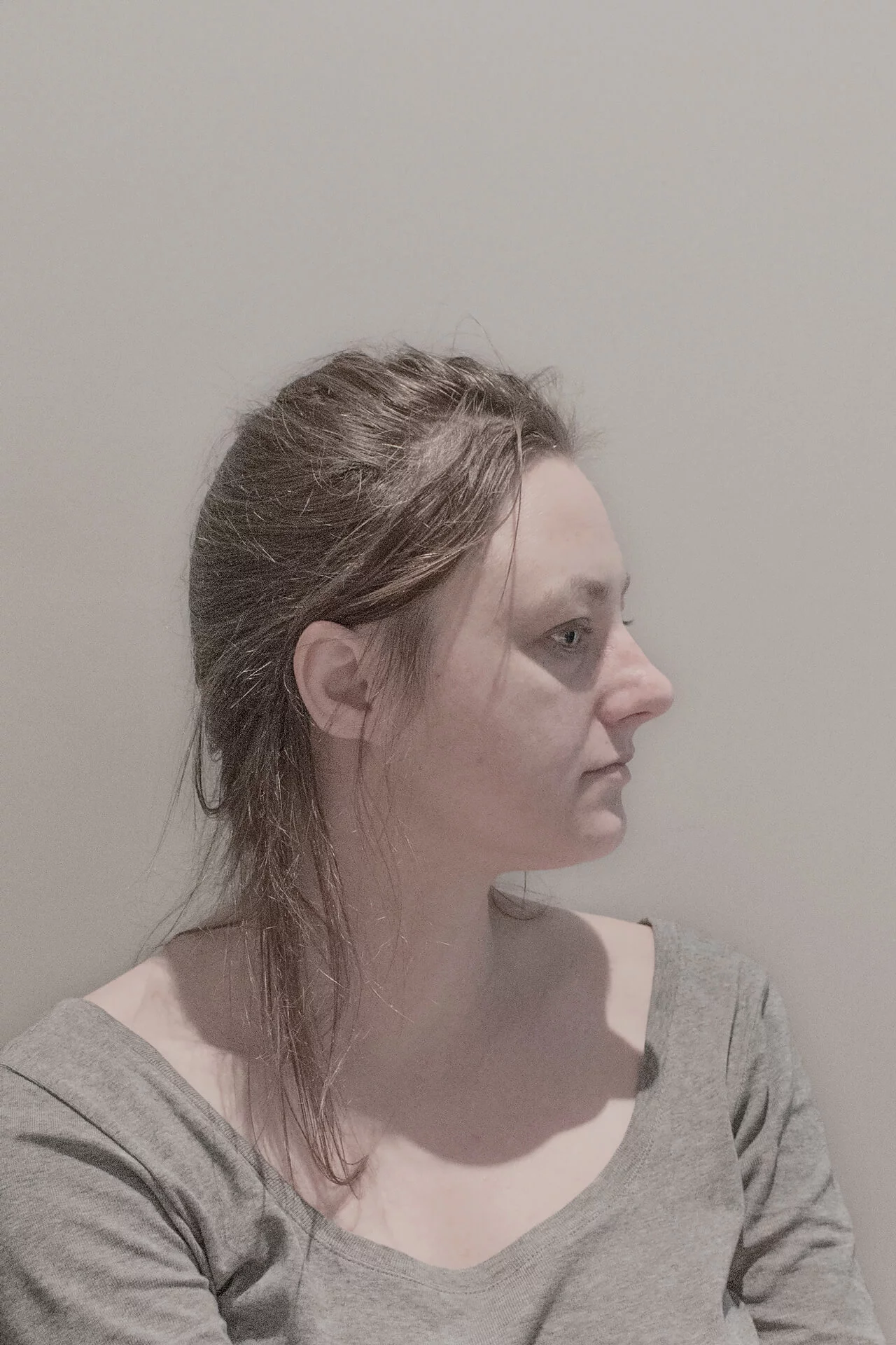 MATTHEW SWARTS A VISUAL portrait of a woman with light skin and brown hair, tied back loosely, sits against a plain light-colored wall, looking to the right. She wears a gray wide-neck top and has a neutral expression in this ART reminiscent of MATTHEW SWARTS.