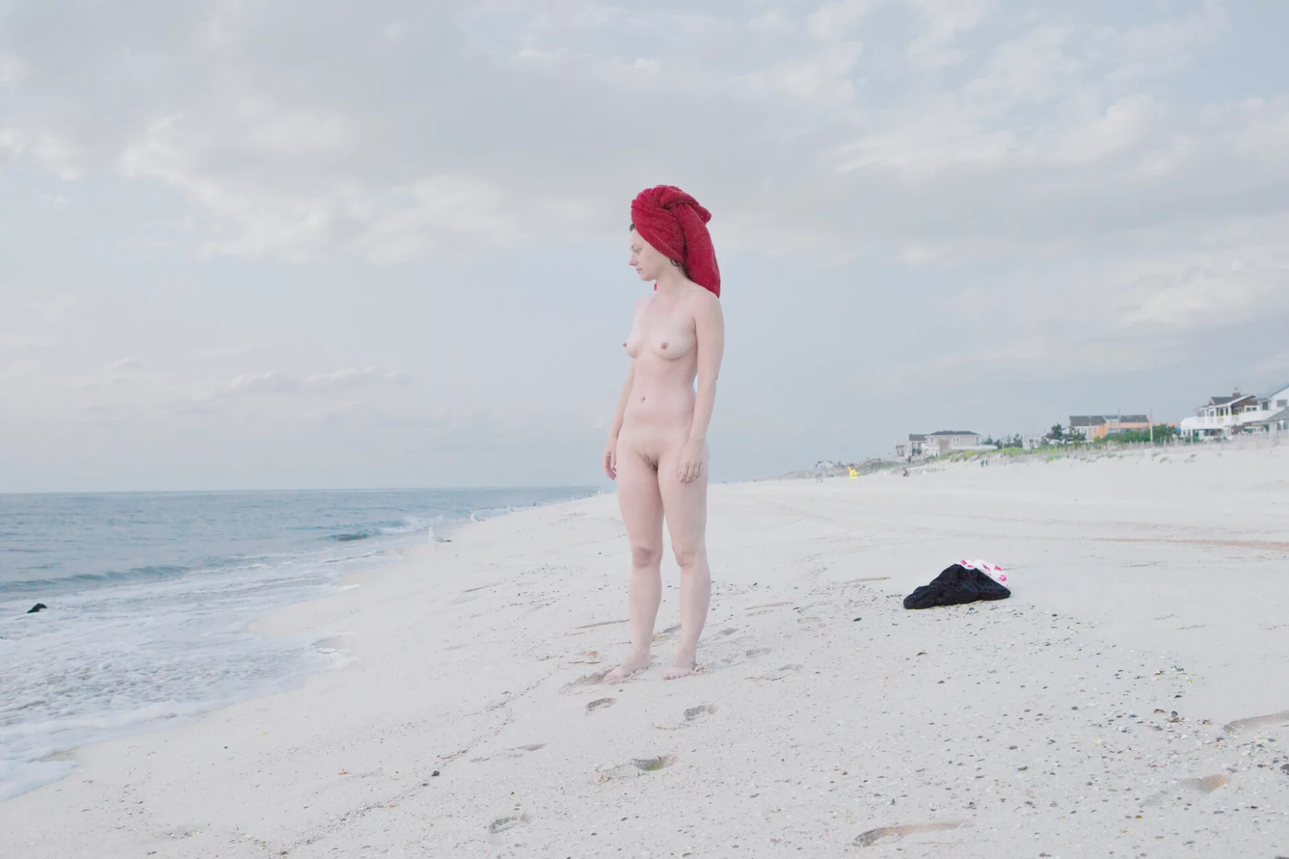 MATTHEW SWARTS A nude person with a red towel wrapped around their head stands on a sandy beach, gazing toward the ocean. Footprints trail behind, and clothes are lying on the sand nearby. The sky is mostly cloudy.