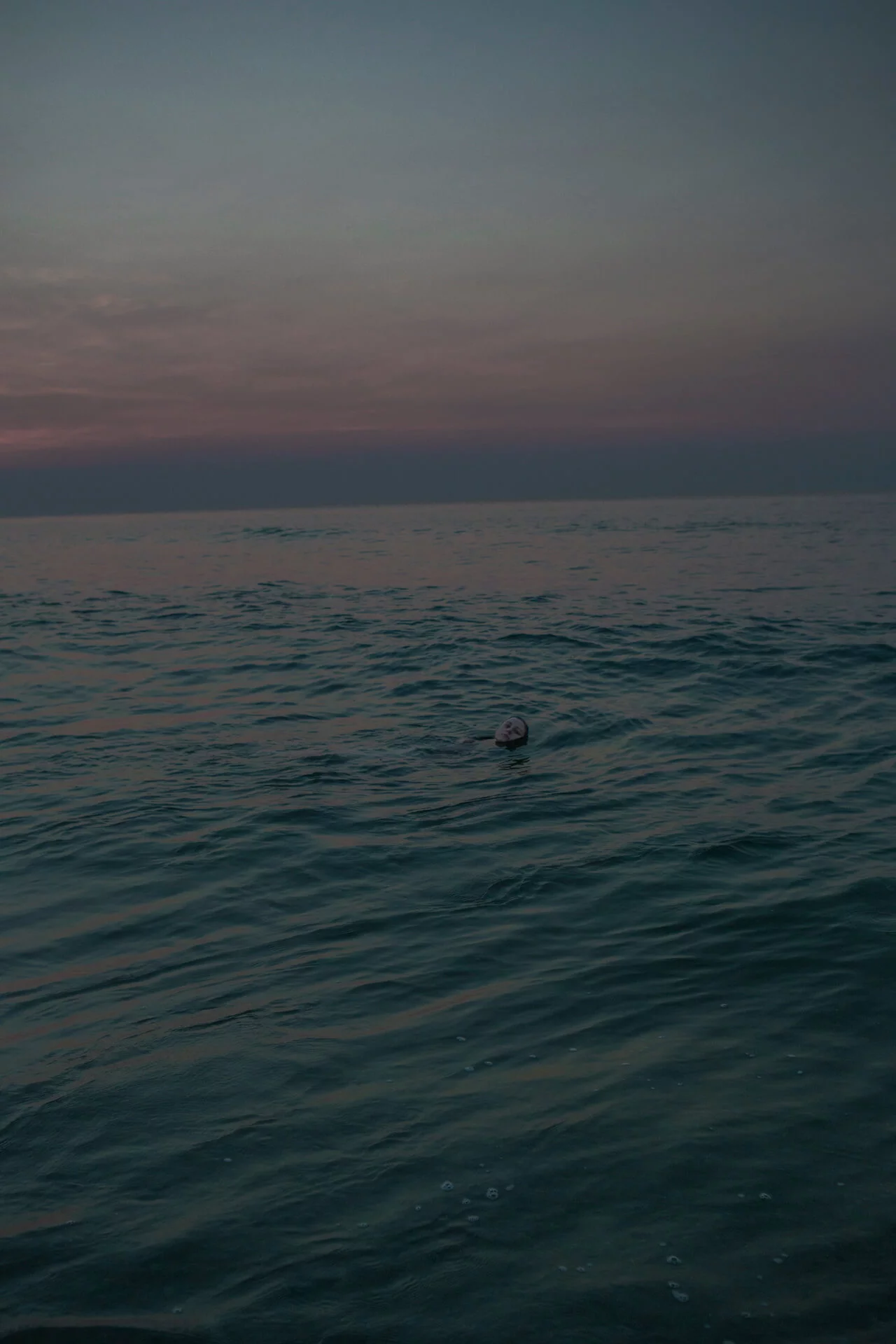 MATTHEW SWARTS A calm, dark ocean at dusk with gentle waves, under a cloudy sky with pink and gray tones. A partially submerged ball floats alone in the water near the center, evoking the contemplative style of ARTIST MATTHEW SWARTS.
