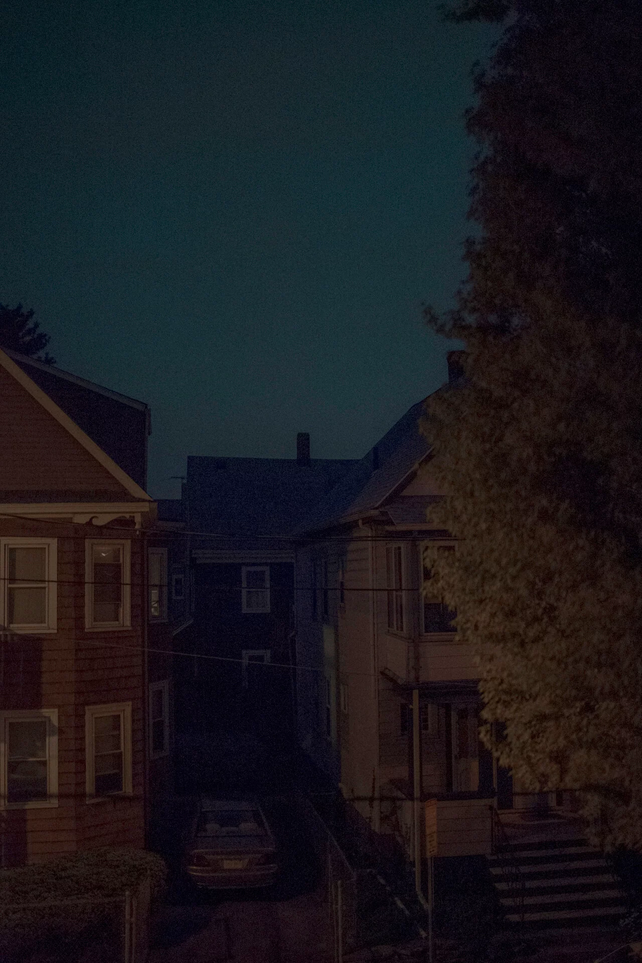 MATTHEW SWARTS A dimly lit residential street at night, captured by photographer Matthew Swarts, shows two houses on either side with a parked car between them. The sky is dark, and one side of a house and a tall tree are faintly illuminated.