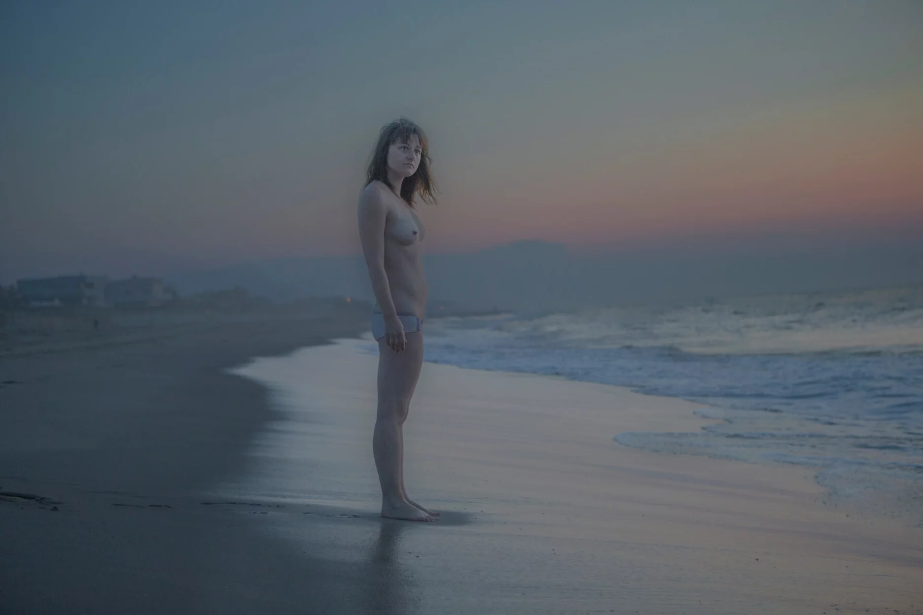 MATTHEW SWARTS A person stands nude on a beach at dusk, looking toward the camera. Gentle waves wash the shore, and the sky is colored with soft hues of blue, pink, and purple as the sun sets. Houses are visible in the distant background.