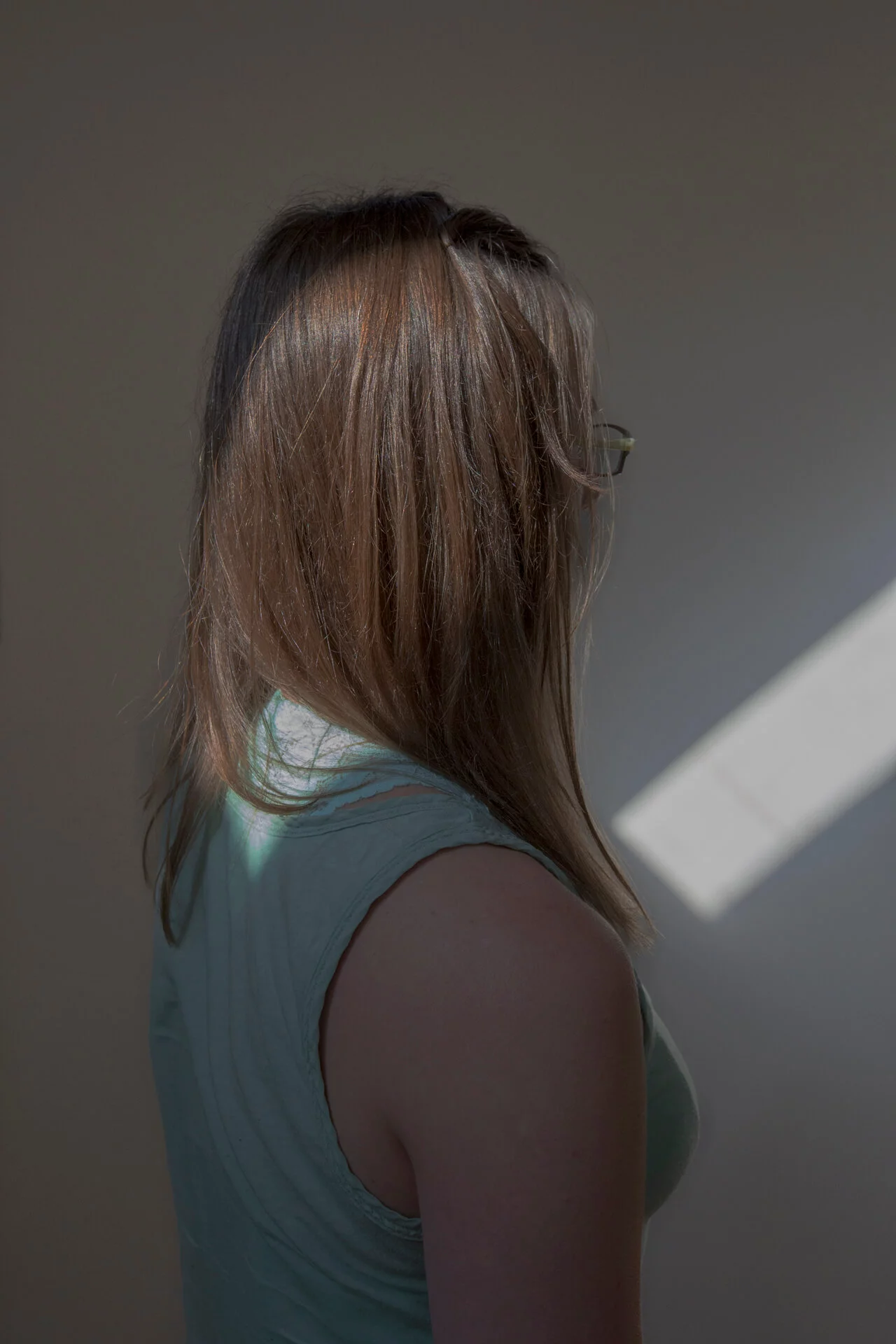 MATTHEW SWARTS A woman with long, light brown hair wearing glasses and a sleeveless light green top stands with her back to the camera, facing a wall lit by a slant of sunlight—a striking visual reminiscent of MATTHEW SWARTS’ art.