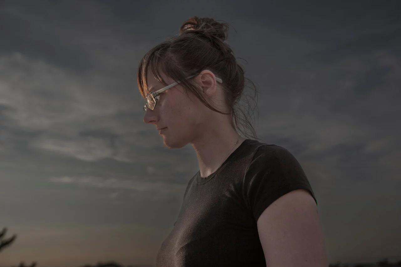 MATTHEW SWARTS A person with glasses and brown hair in a bun stands outdoors at dusk, wearing a dark t-shirt and looking down, set against a contemporary, cloudy sky in the background—evoking the visual style of Matthew Swarts.