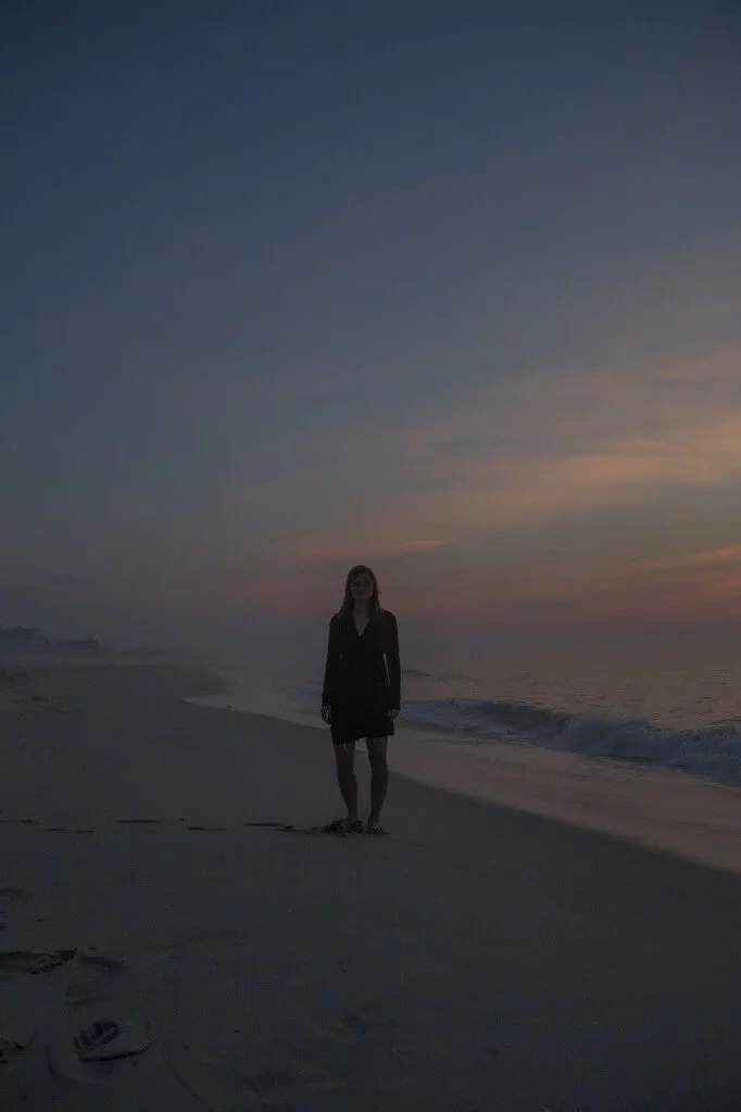 MATTHEW SWARTS A photographer in a dark outfit stands on a quiet beach at sunset, capturing gentle waves and a colorful sky—turning the scene into art.