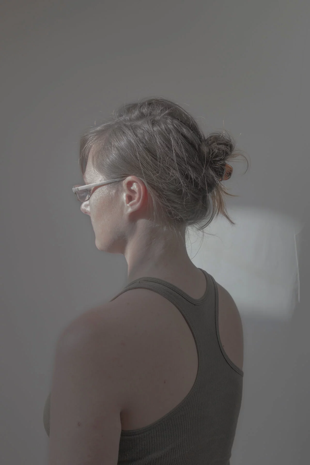 MATTHEW SWARTS A woman with brown hair in a messy bun, wearing glasses and a dark tank top, stands facing away from the camera in soft, natural light against a plain background—a contemporary visual captured by the photographer.