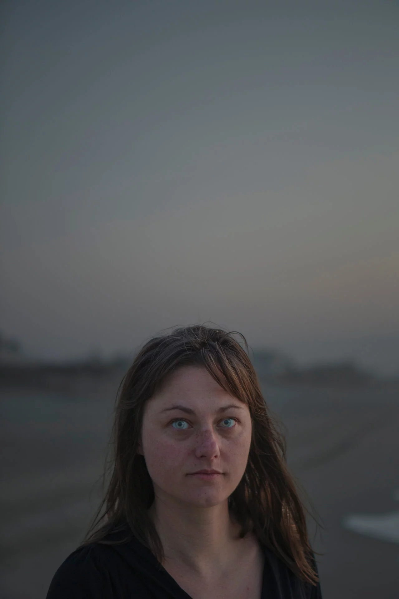MATTHEW SWARTS Close-up portrait of a woman with bright blue eyes looking at the camera. The background is a blurry, hazy landscape.