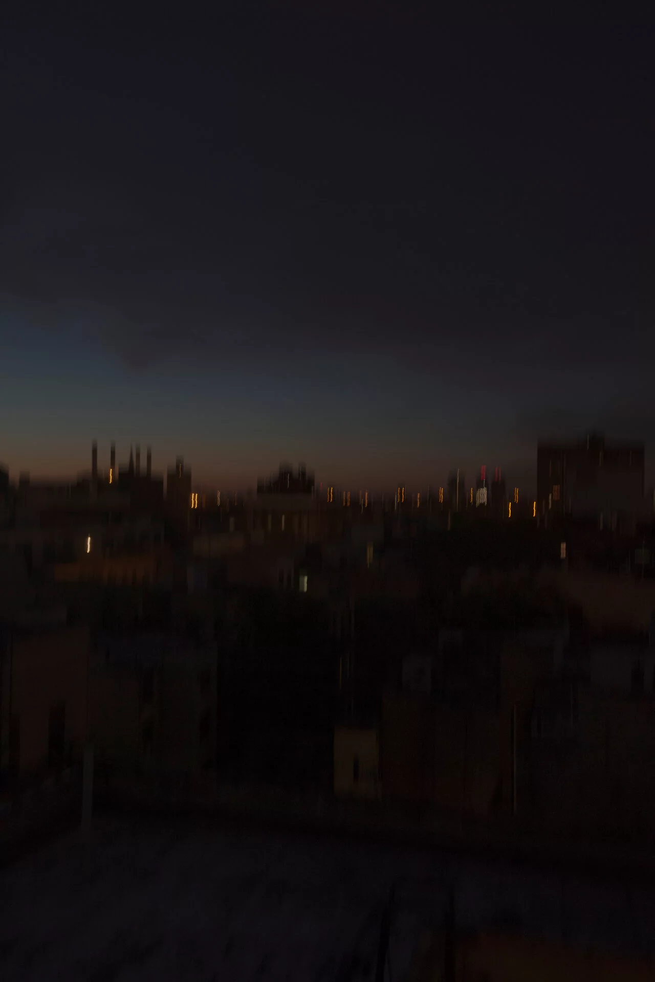 MATTHEW SWARTS A dark cityscape at dusk with blurred buildings in the foreground and scattered lights dotting the horizon under a dim, cloudy sky captures the moody vision of a contemporary photographer.