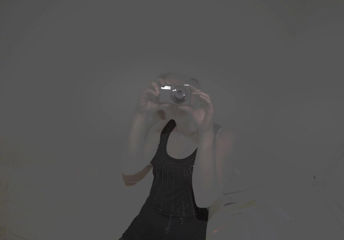 MATTHEW SWARTS A VISUAL ARTIST wearing a sleeveless black top takes a photo of their reflection in a dimly lit mirror, camera held in front of their face, creating an image that is slightly obscured—reminiscent of MATTHEW SWARTS' introspective style.