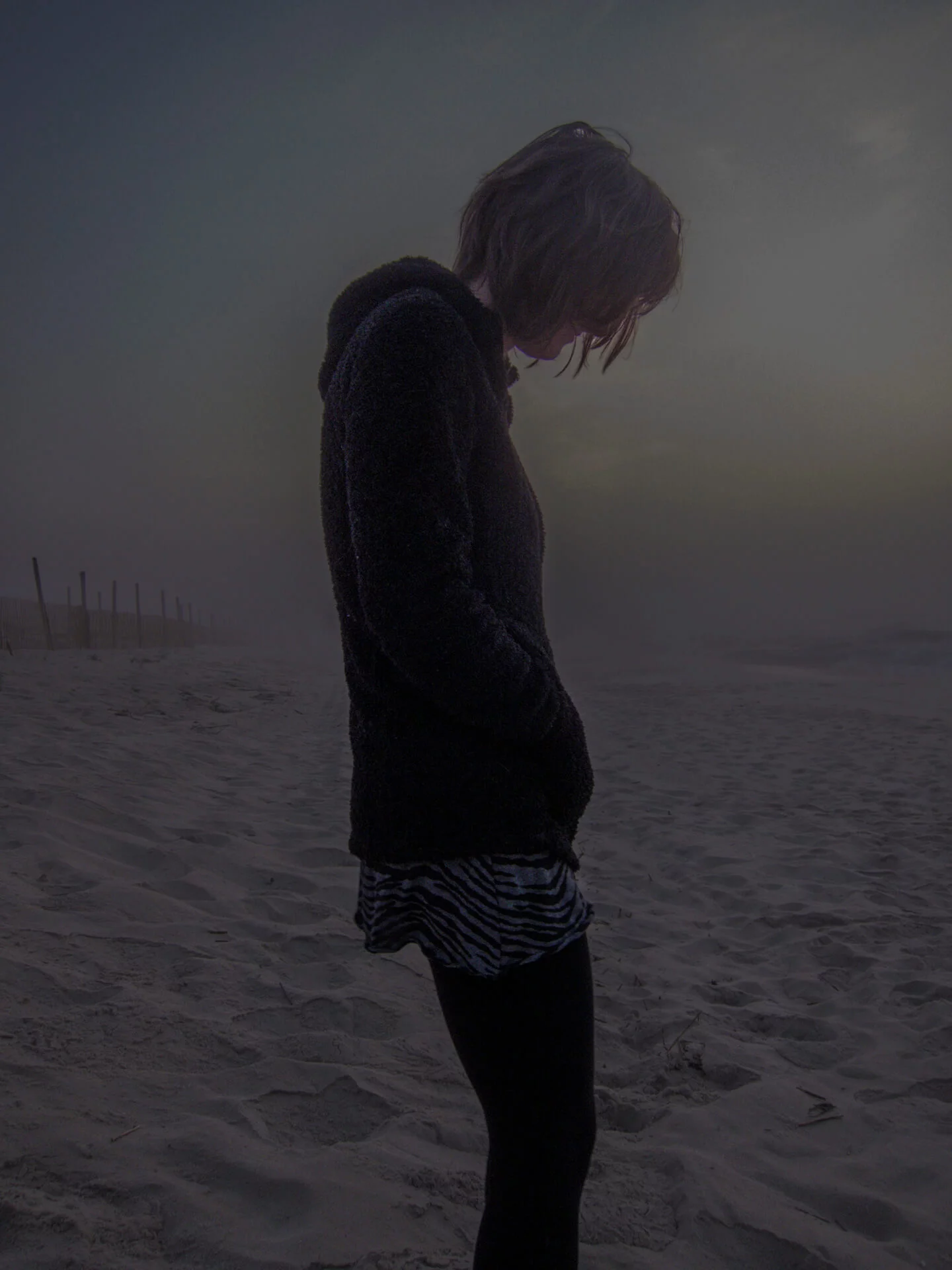 MATTHEW SWARTS A person stands on a sandy beach at dusk, captured by a photographer in a moment of quiet reflection. Their dark hoodie and striped skirt evoke contemporary art against the dim, overcast sky with a faint glow on the horizon.