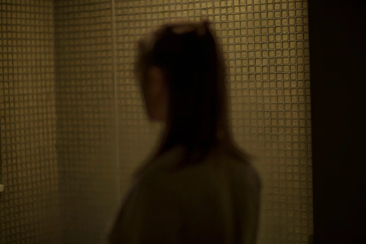 MATTHEW SWARTS A blurry silhouette of a person with long hair stands in front of a wall covered in small square tiles, lit by soft, warm light—an evocative scene that could inspire any artist or photographer.