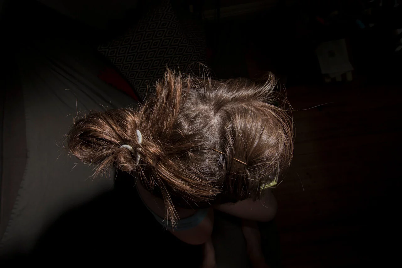 MATTHEW SWARTS A contemporary visual artist with brown hair tied in a messy bun sits on a chair, viewed from above. The background features part of a patterned pillow and a wooden floor, partially covered in shadow.