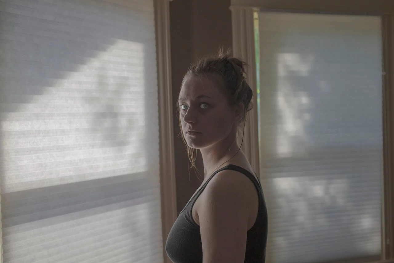 MATTHEW SWARTS A woman with light skin and tied-back hair stands in a dimly lit room, wearing a black tank top, looking towards the camera as soft sunlight filters through window blinds—a striking visual reminiscent of Matthew Swarts’ art.
