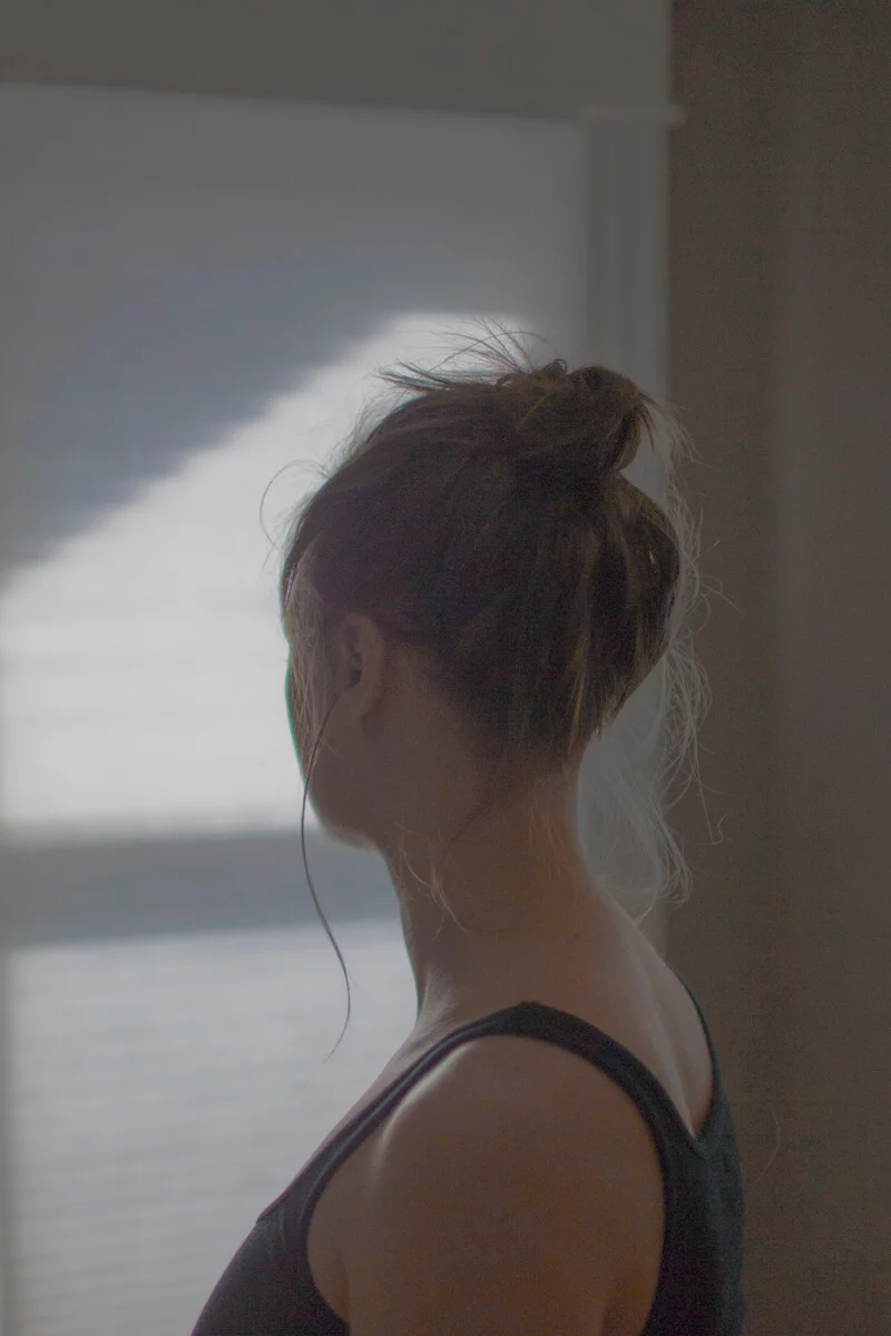 MATTHEW SWARTS A woman with brown hair in a messy bun stands indoors, facing a window with blinds. Soft light casts visual shadows on the wall, illuminating her neck and shoulder as she looks away from the camera, evoking the art of MATTHEW SWARTS.